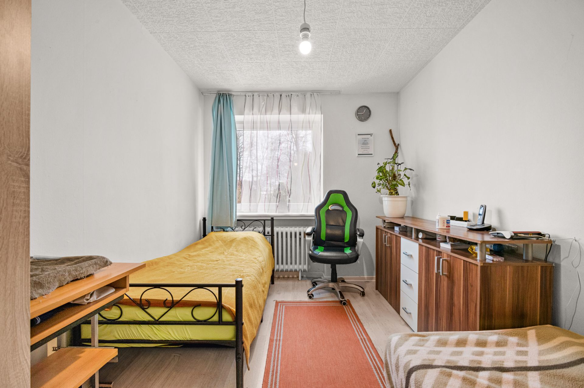 Ein einfach eingerichtetes Schlafzimmer mit Bett, Schreibtisch, Stuhl, Kommode und Fenster mit Vorhängen.