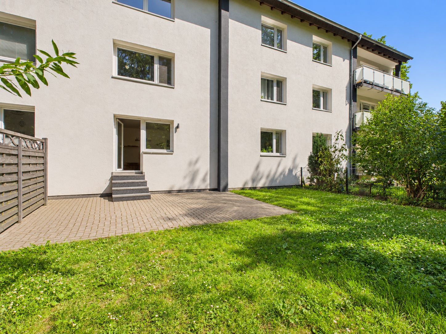 Hinterhof eines Wohnhauses mit Terrasse, Rasenfläche und mehreren Fenstern.