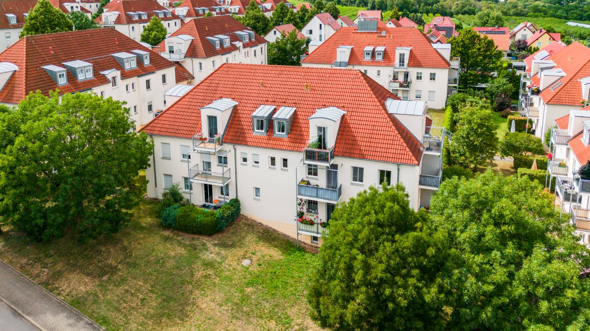 Mehrfamilienhaus mit rotem Dach und mehreren Balkonen, umgeben von Bäumen.