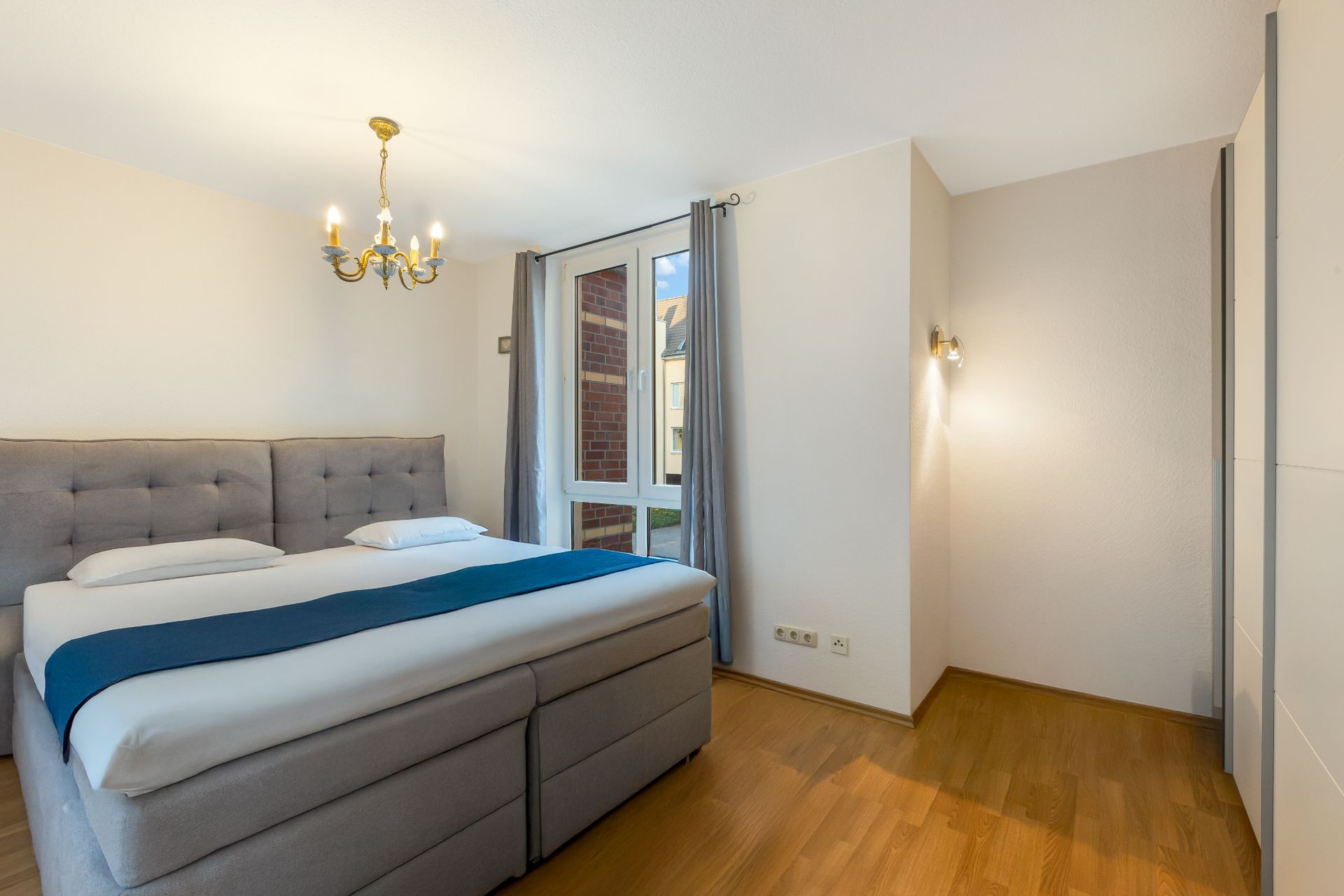 Schlafzimmer mit Doppelbett, Holzfußboden, Fenster mit Vorhängen und Kronleuchter.