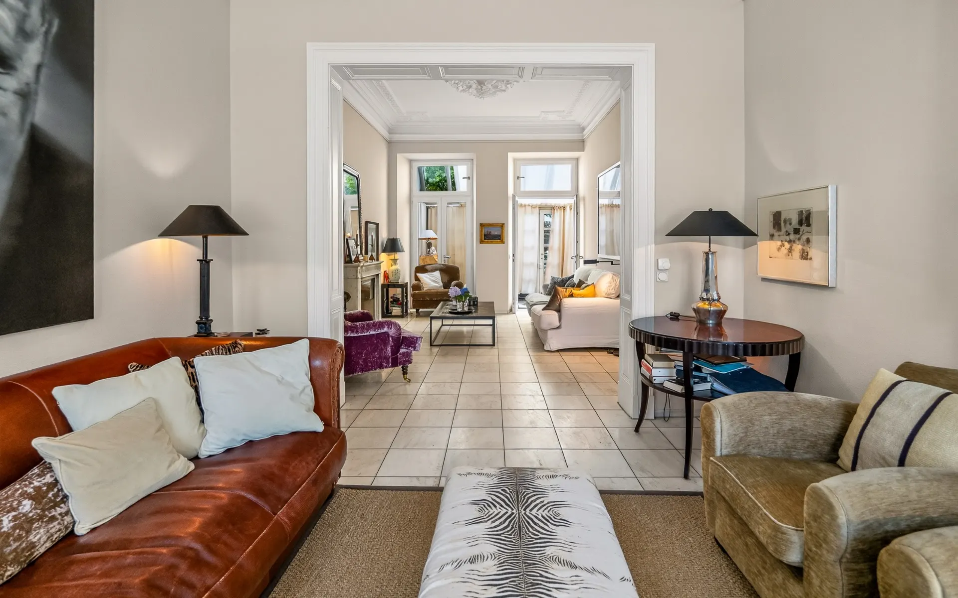 Helles Wohnzimmer mit Ledersofa, Sesseln, Tischlampe und Blick in einen zweiten Raum mit Sofas.