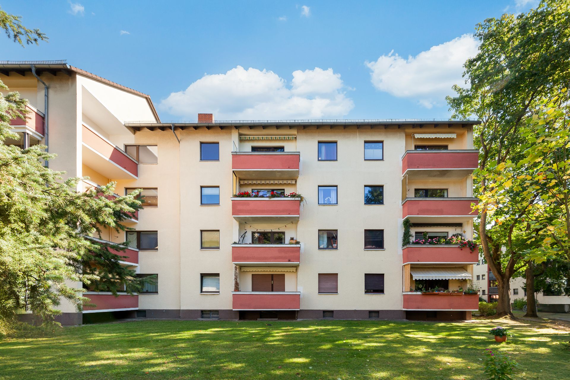 Vierstöckiges Wohnhaus mit Balkonen und Rasenfläche, umgeben von Bäumen.