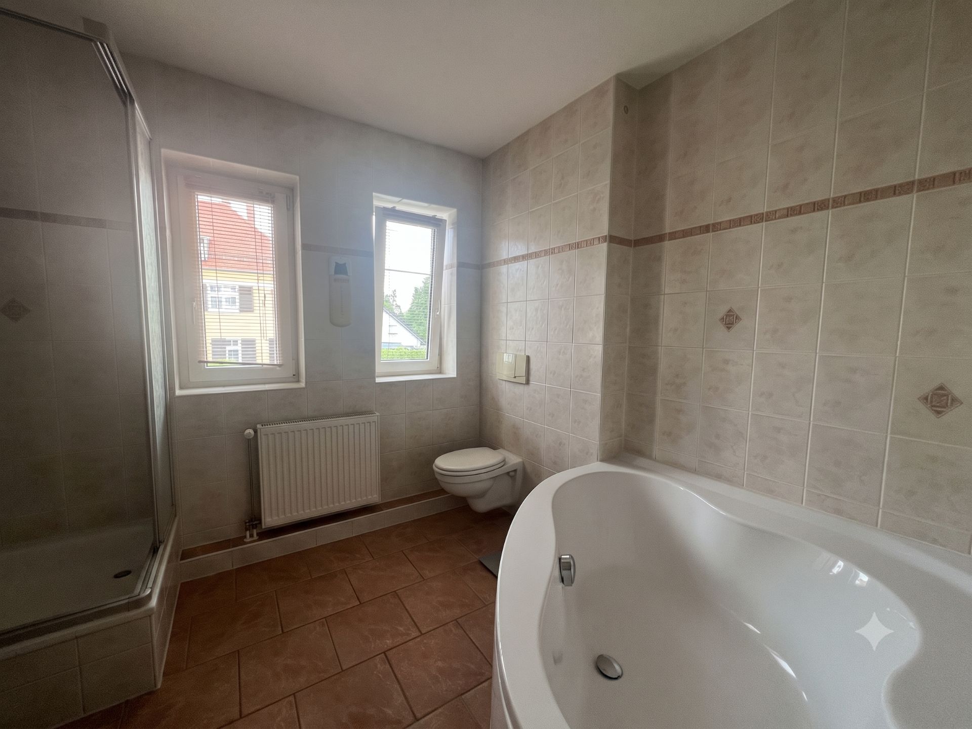 Helles Badezimmer mit Dusche, Badewanne, Toilette und zwei Fenstern.