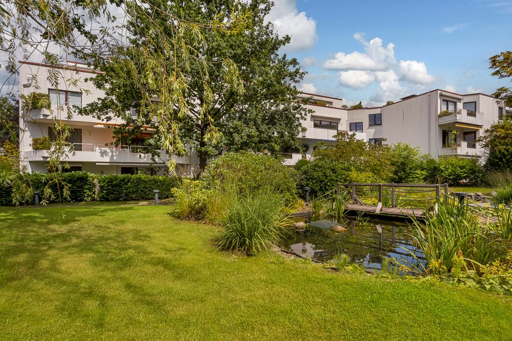 Moderne Wohngebäude mit Balkonen, Garten, Teich und kleiner Holzbrücke im Vordergrund.
