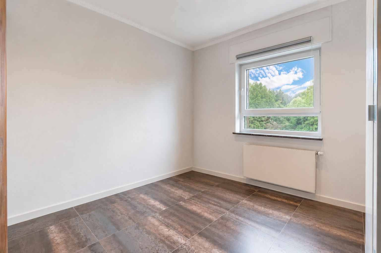 Leerer Raum mit braunem Fliesenboden, weißer Wand und Fenster mit Blick ins Grüne.