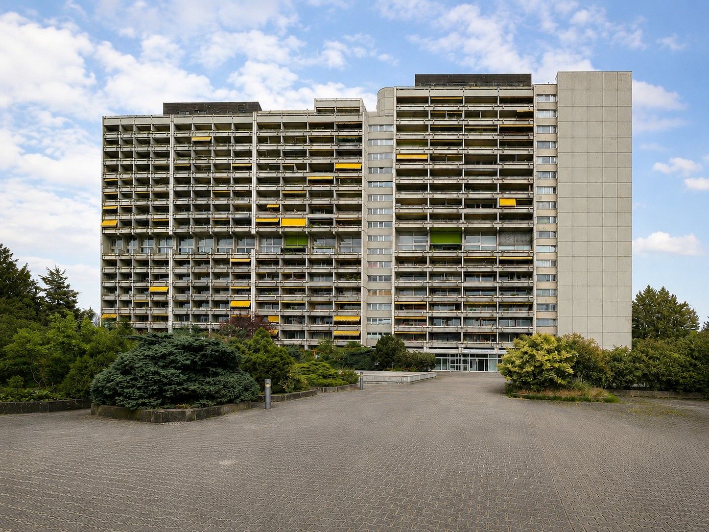 Wohngebäude mit Balkonen und bunten Markisen, davor Gehweg und Sträucher.