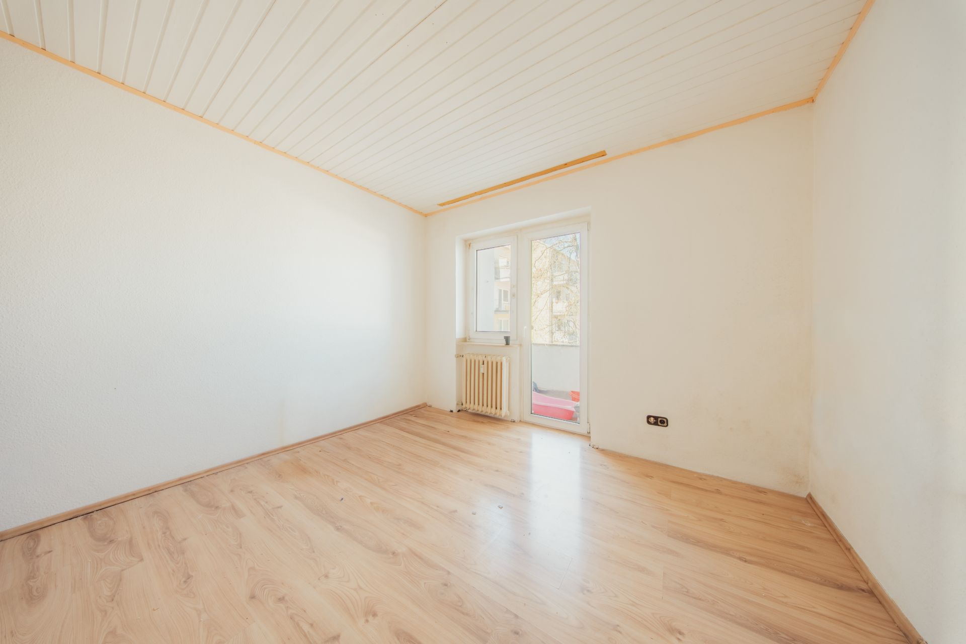 Leerer Raum mit hellem Holzboden, weißen Wänden und Tür zu einem Balkon.