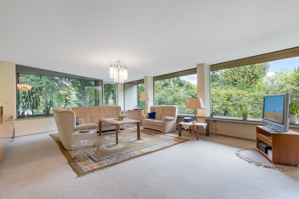 Helles Wohnzimmer mit cremefarbenen Sofas, großem Teppich, TV und breiten Fenstern mit Blick ins Grüne.