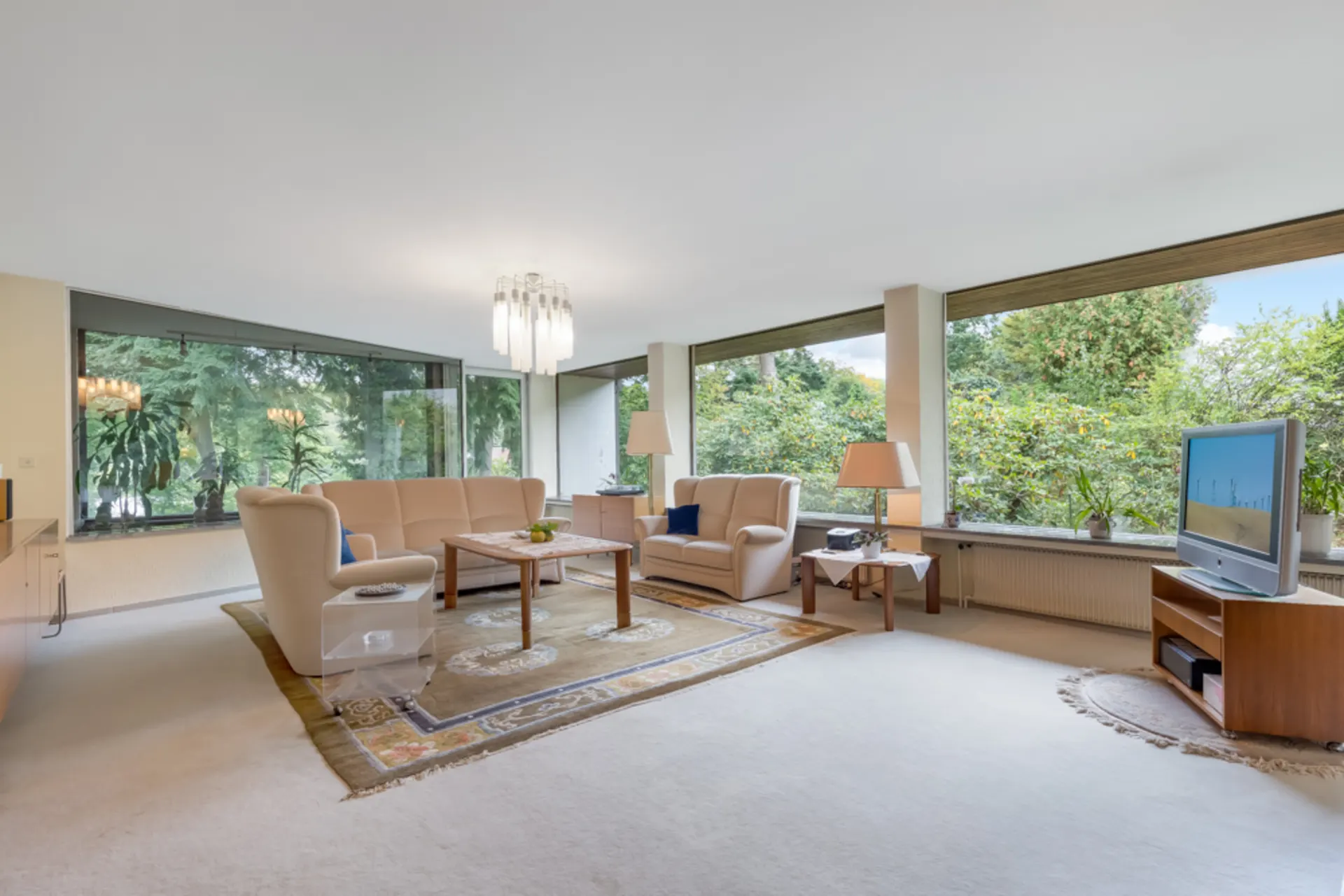 Helles Wohnzimmer mit cremefarbenen Sofas, großem Teppich, TV und breiten Fenstern mit Blick ins Grüne.