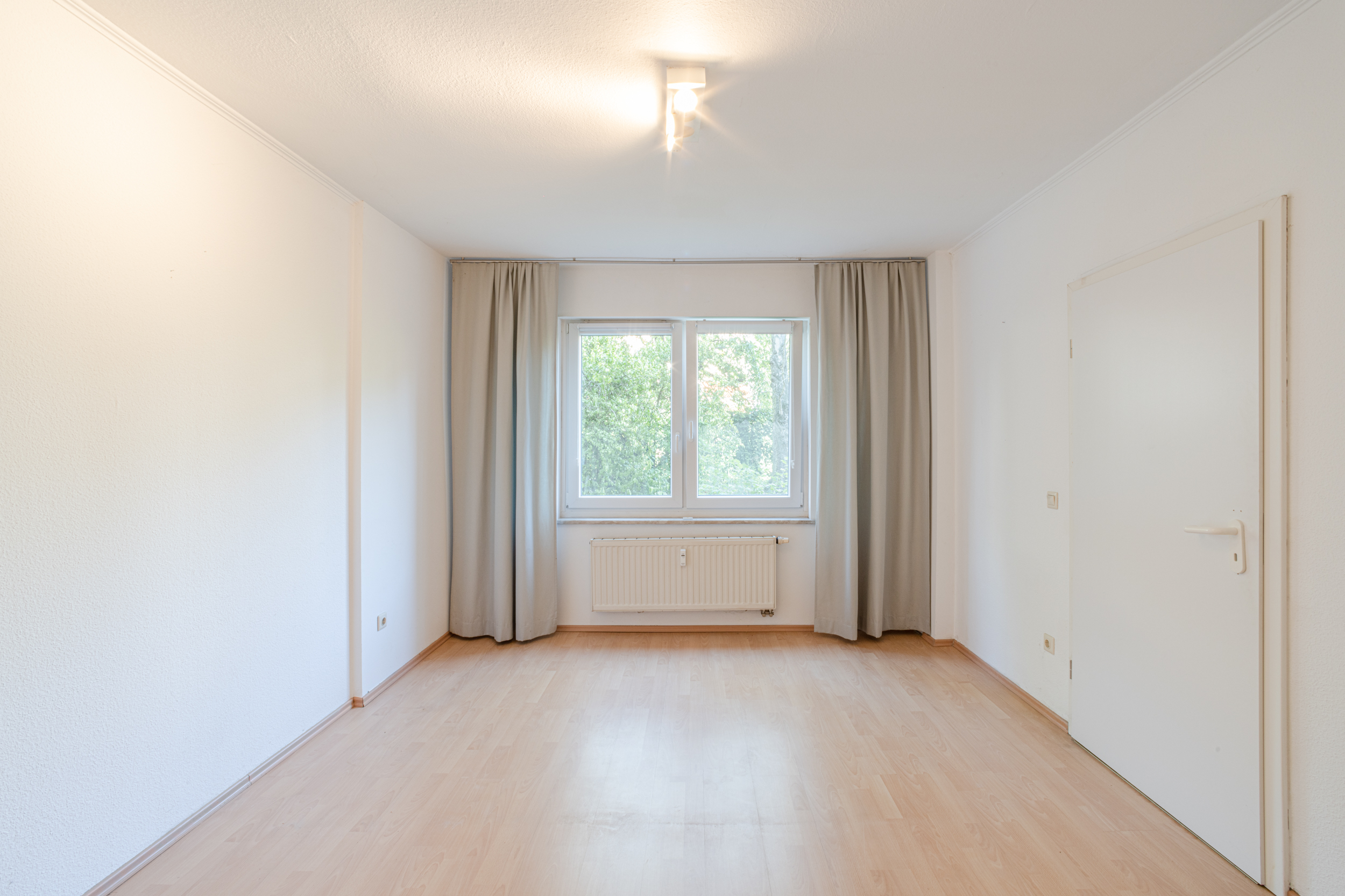Helles, leeres Zimmer mit Holzfußboden, Fenster mit Vorhängen und weißer Tür.
