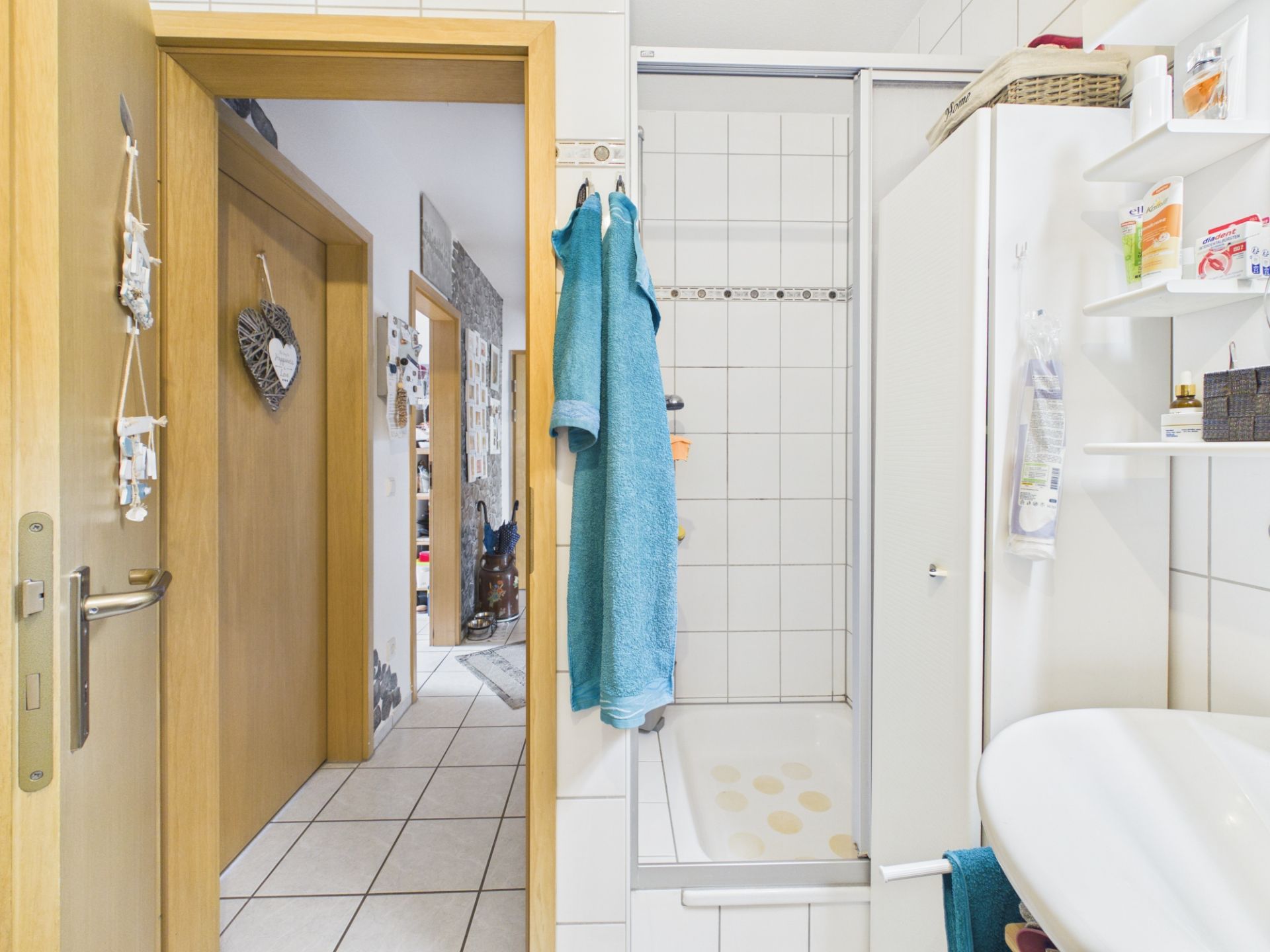 Helles Badezimmer mit Dusche, Waschbecken, Handtüchern und Blick in den Flur einer Wohnung.