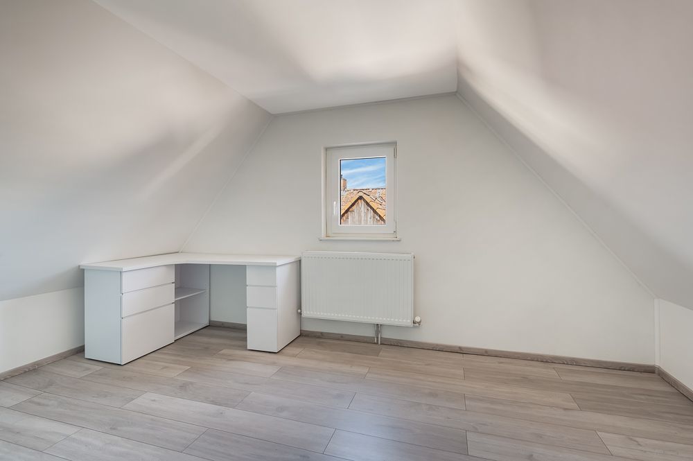 Helles Dachzimmer mit Schreibtisch, Fenster und Heizkörper, auf hellem Holzboden.