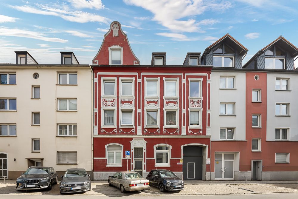 Mehrfamilienhaus mit roter Fassade und weißen Zierleisten, drei Etagen, Autos parken davor.