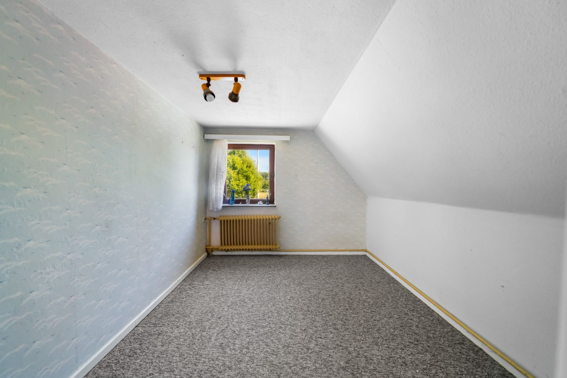 Leeres Zimmer mit schräger Decke, Teppichboden, Fenster und Heizkörper.