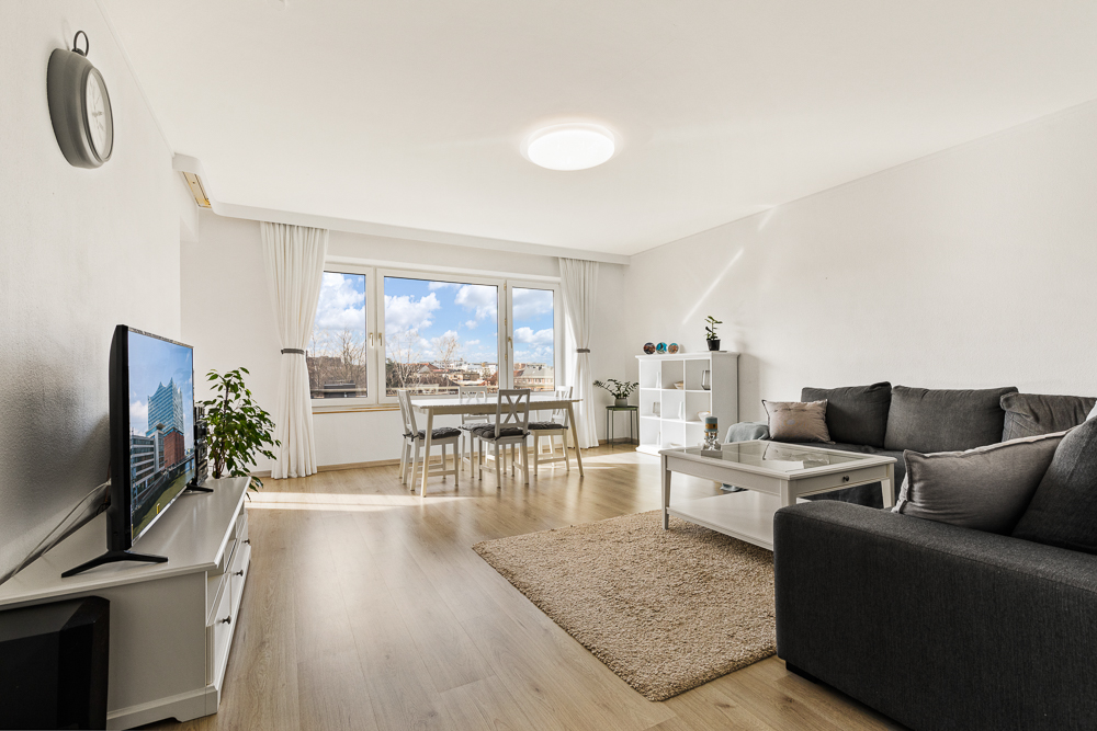 Helles Wohnzimmer mit großem Fenster, grauem Sofa, TV, Esstisch und Regal auf Holzfußboden.