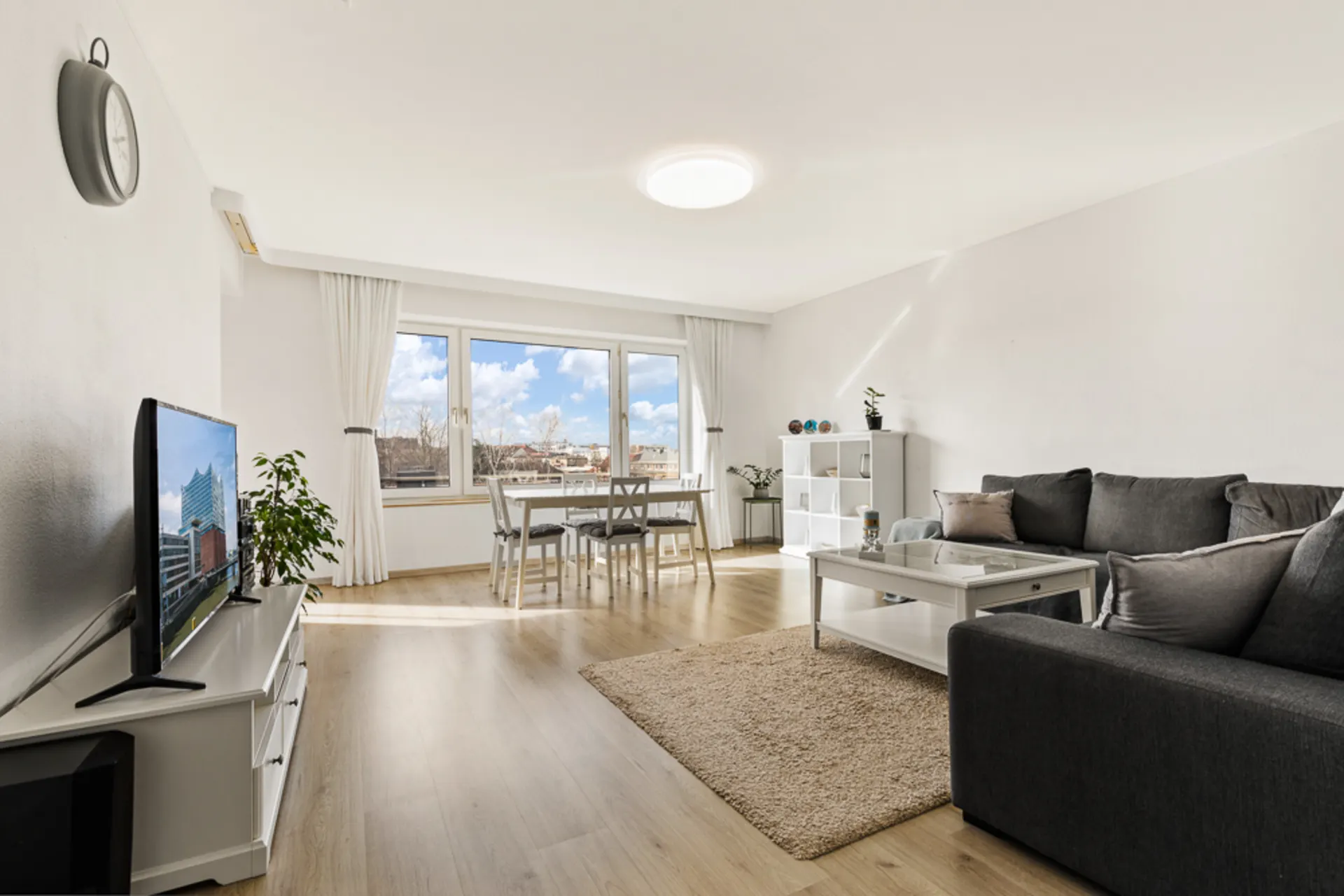 Helles Wohnzimmer mit großem Fenster, grauem Sofa, TV, Esstisch und Regal auf Holzfußboden.