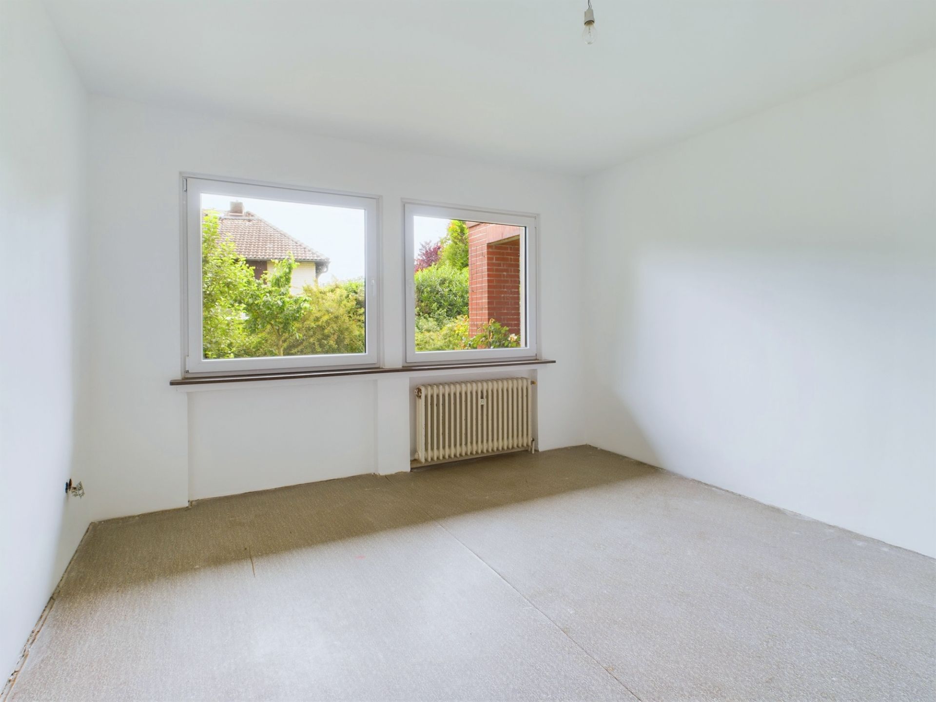 Leerer, heller Raum mit zwei großen Fenstern und einem Heizkörper unter der Fensterbank.