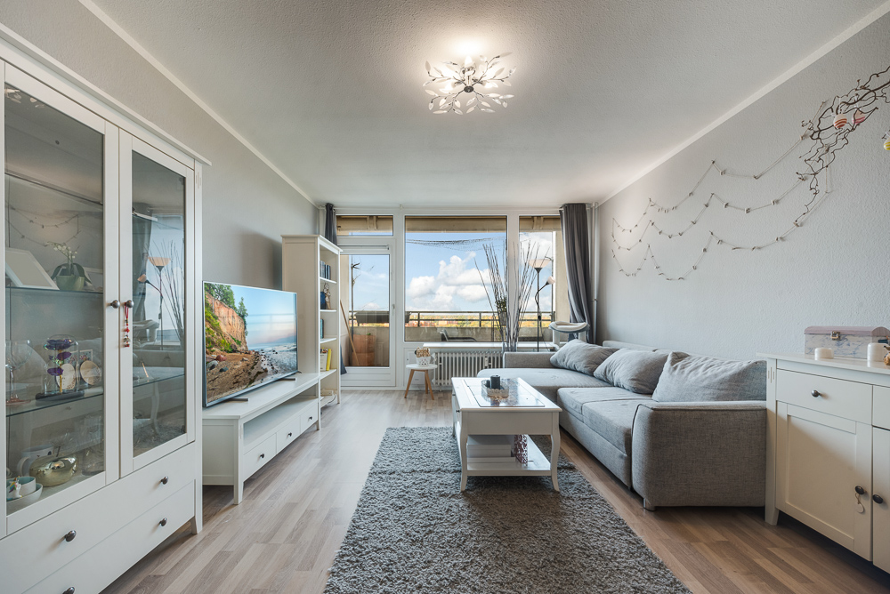 Helles Wohnzimmer mit grauem Sofa, Teppich, Fernseher und großem Fenster mit Blick nach draußen.