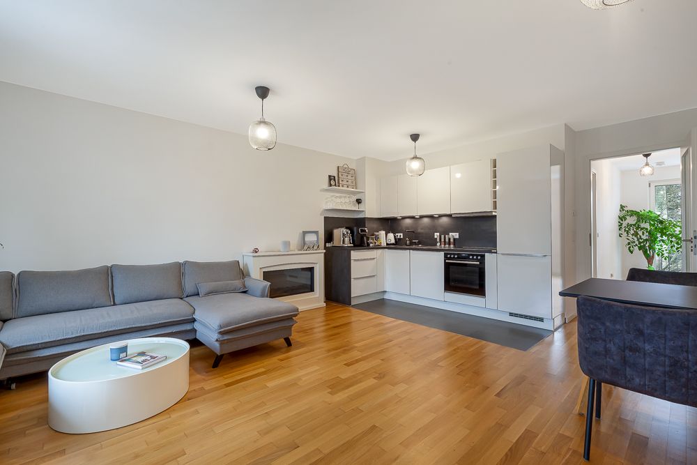 Helles Wohnzimmer mit grauem Sofa, offener weißer Küche und Esstisch auf Parkettboden.