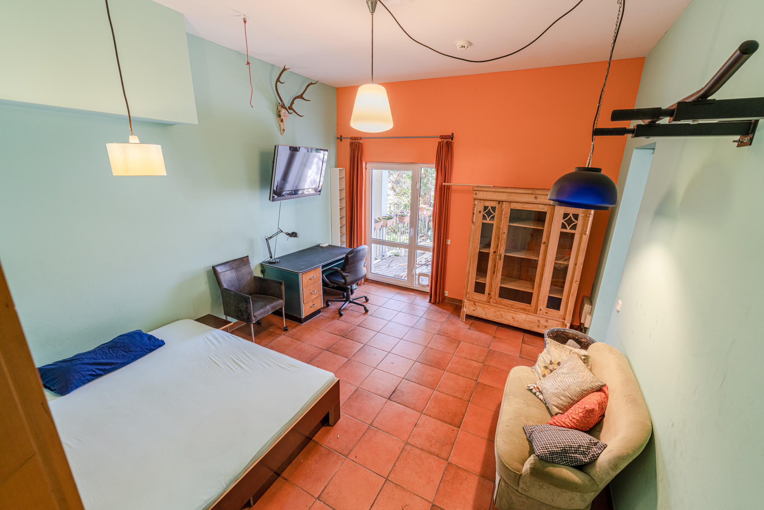 Zimmer mit Bett, Schreibtisch, Sessel, Sofa und großer Vitrine, Zugang zu Balkon durch Glastür.