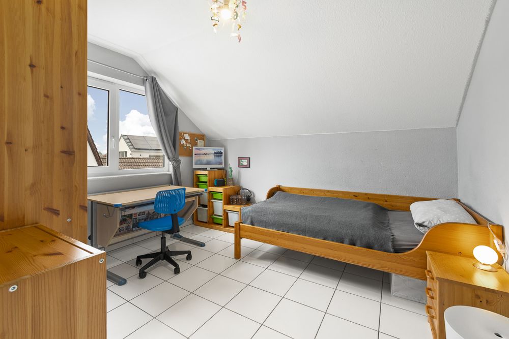 Helles Schlafzimmer mit Einzelbett, Schreibtisch am Fenster und weißem Fliesenboden.