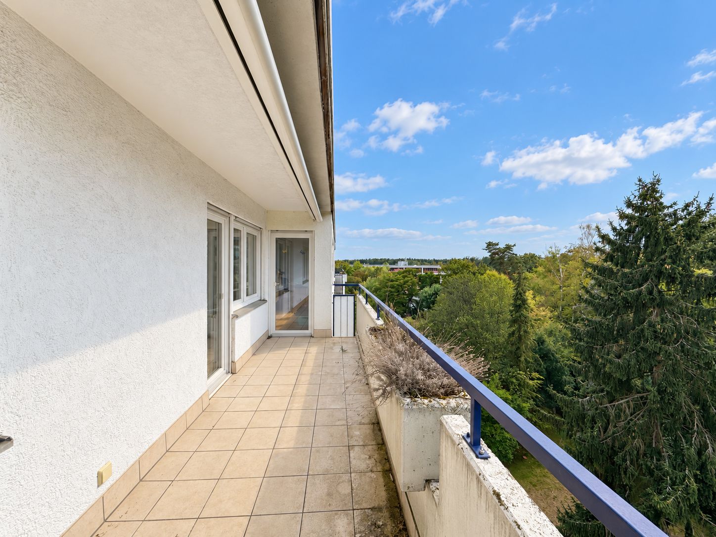 Heller Balkon mit Fliesenboden und Glasgeländer, Ausblick auf Bäume und blauen Himmel.