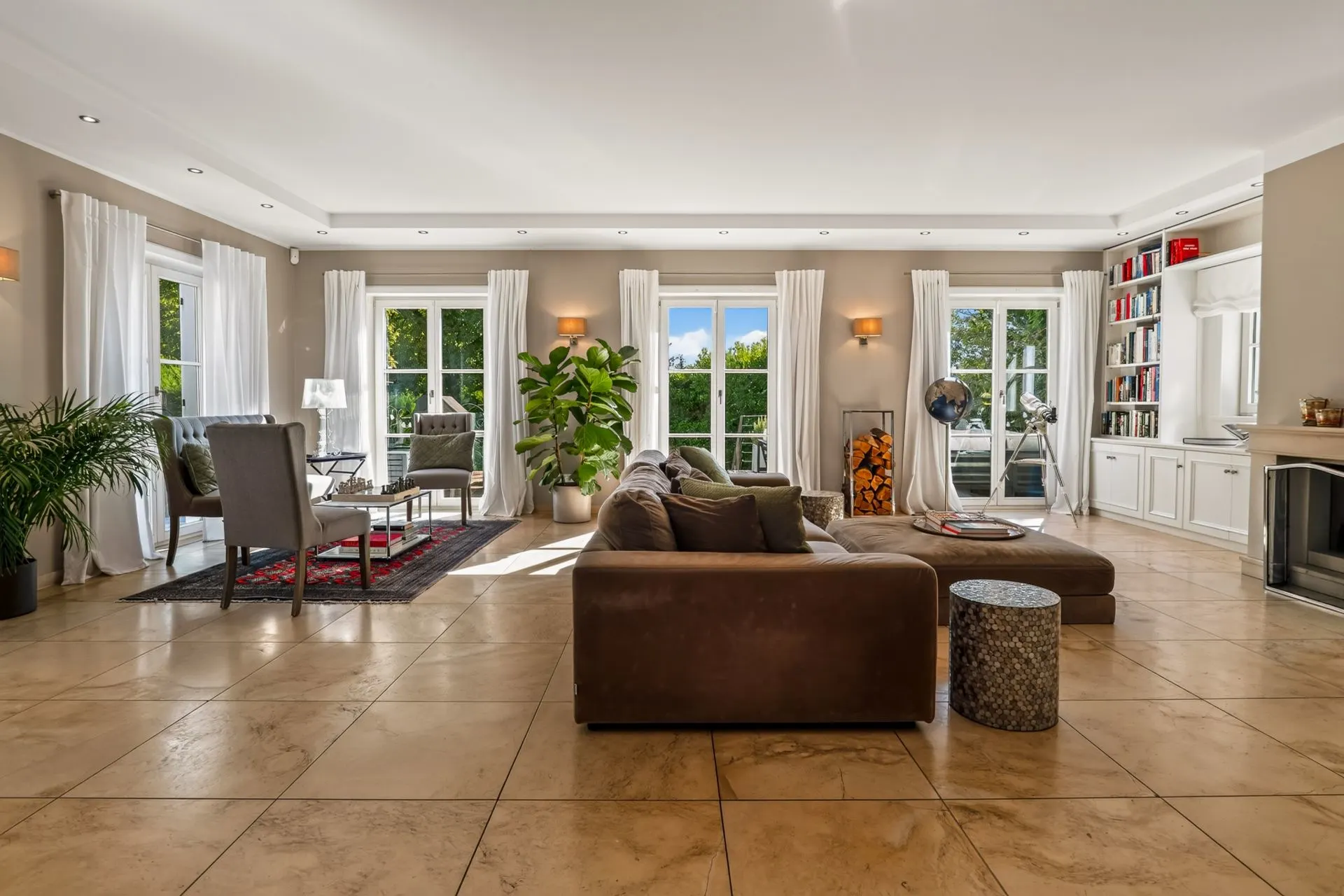 Helles Wohnzimmer mit braunem Sofa, Sesseln, großen Fenstern, Pflanzen und Bücherregal.