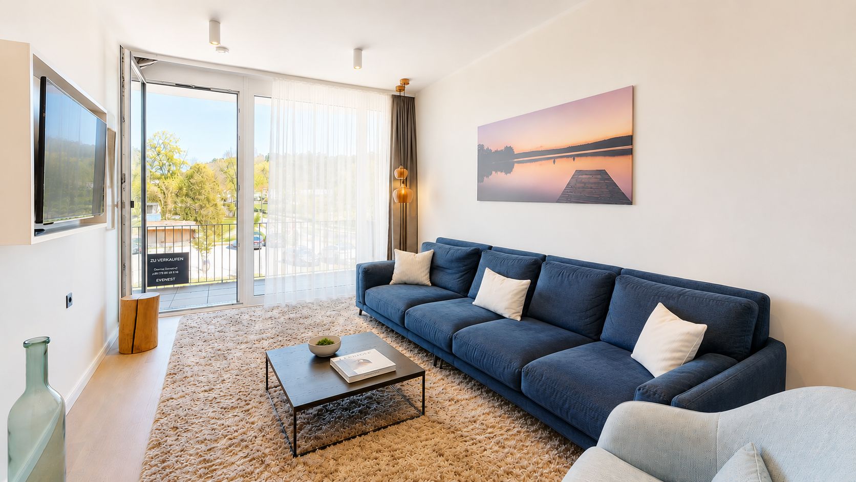 Helles Wohnzimmer mit blauem Sofa, Teppich, Couchtisch und großem Fenster mit Vorhängen.