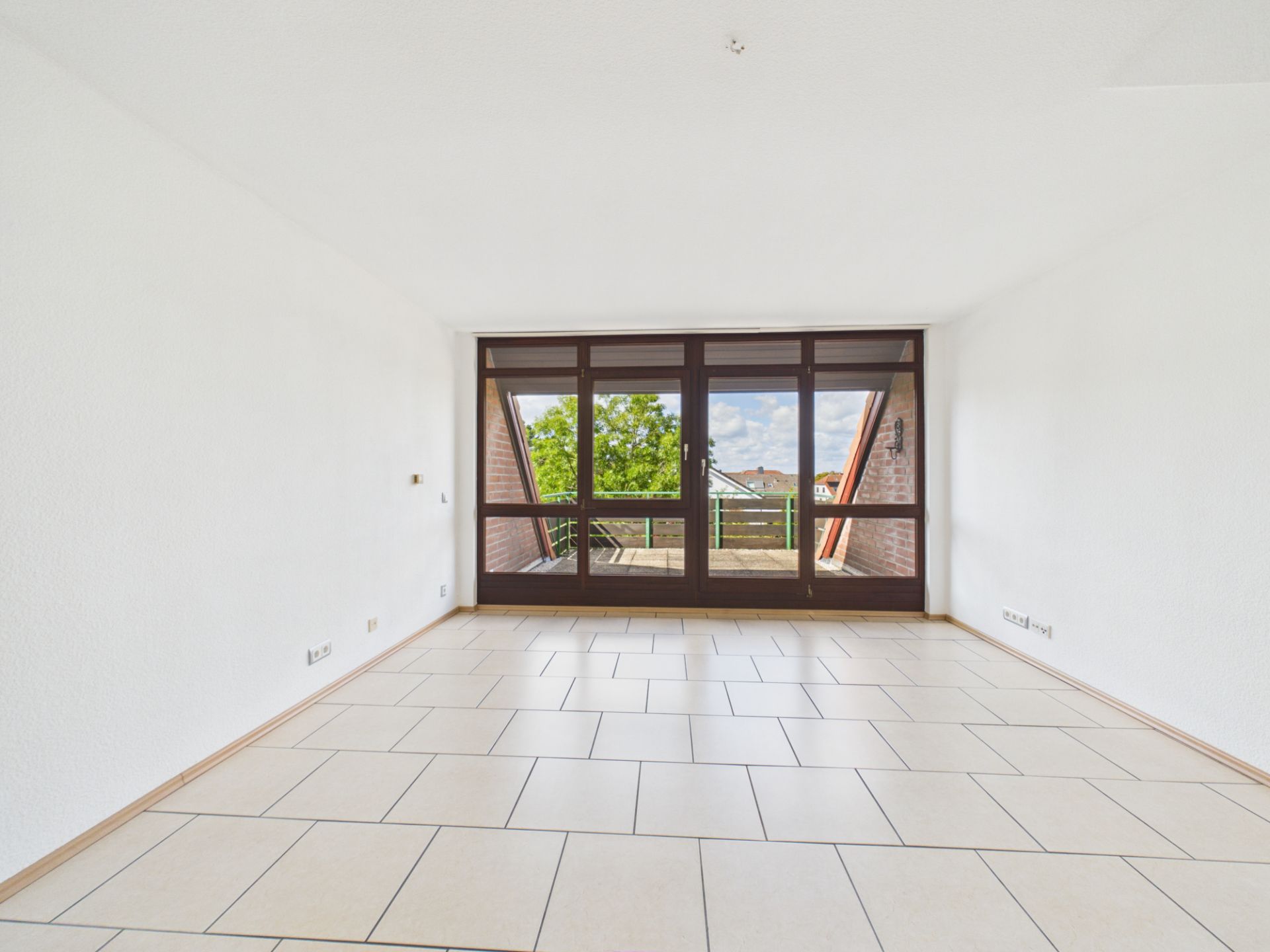 Leeres, helles Zimmer mit Fliesenboden und großer Fensterfront zu einem Balkon.