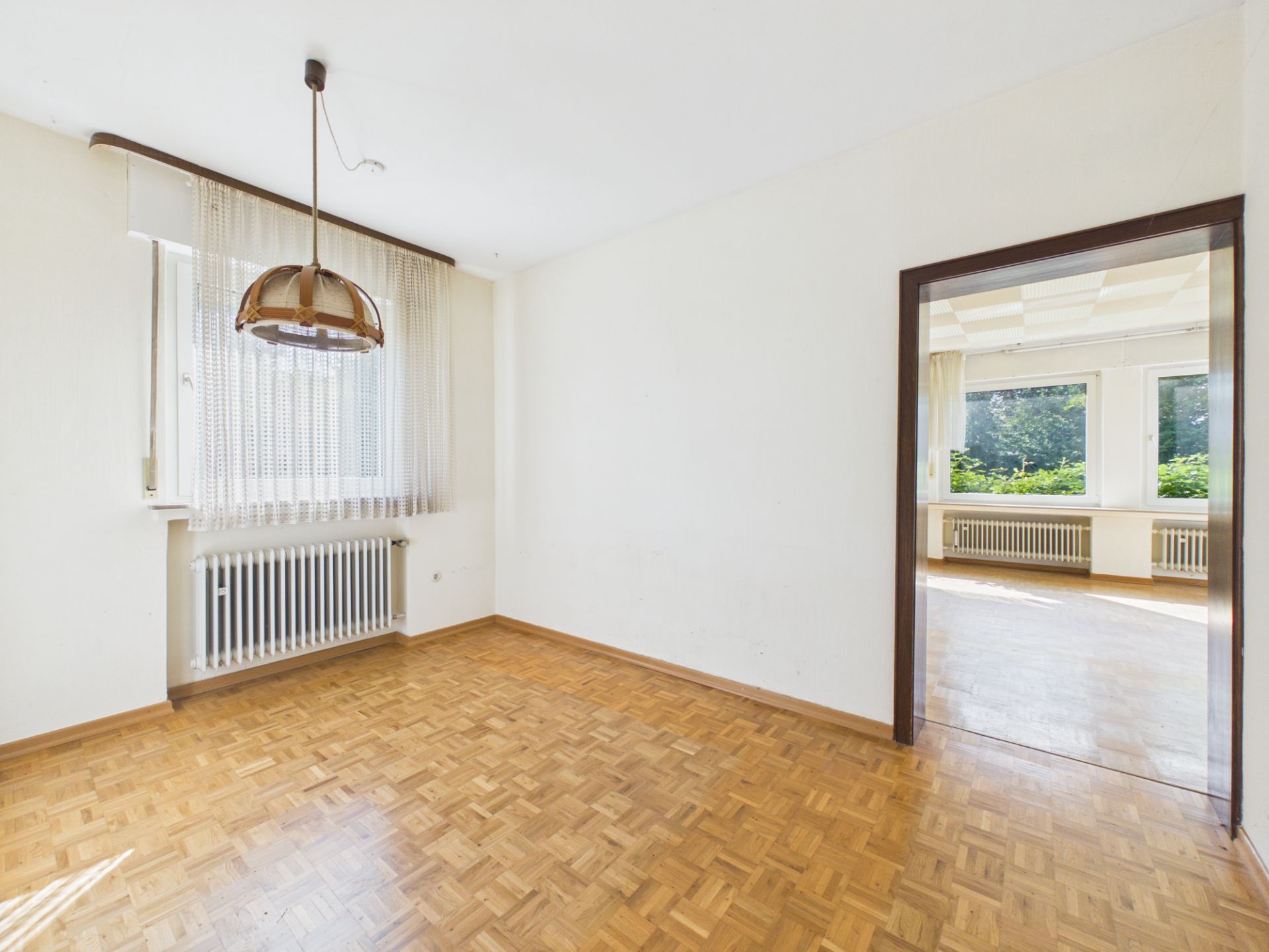 Heller, leerer Raum mit Parkett, Fenster, Heizkörper und offener Tür zu einem weiteren Zimmer.