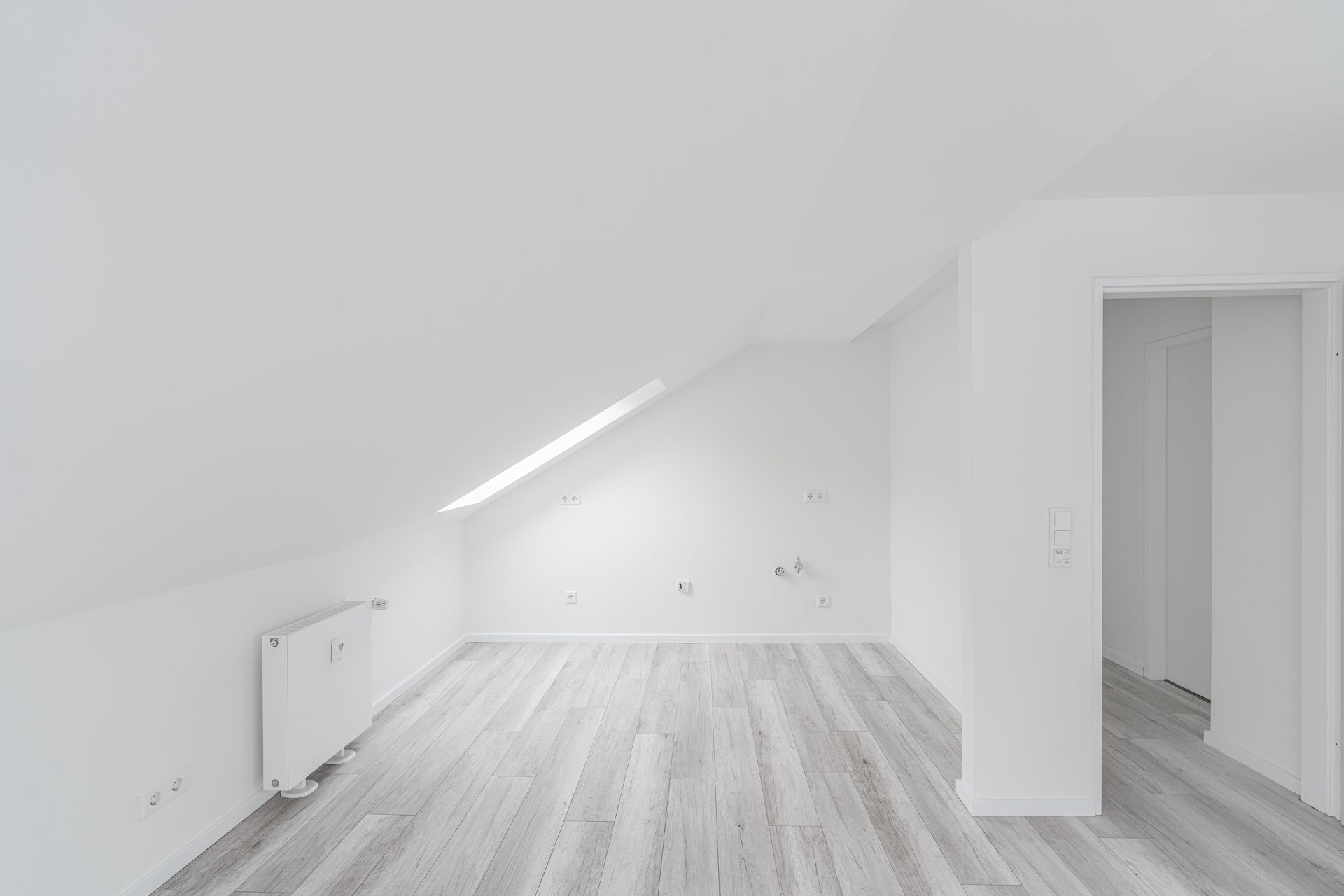 Leerer Raum mit Dachschräge, großem Fenster und hellem Holzboden.