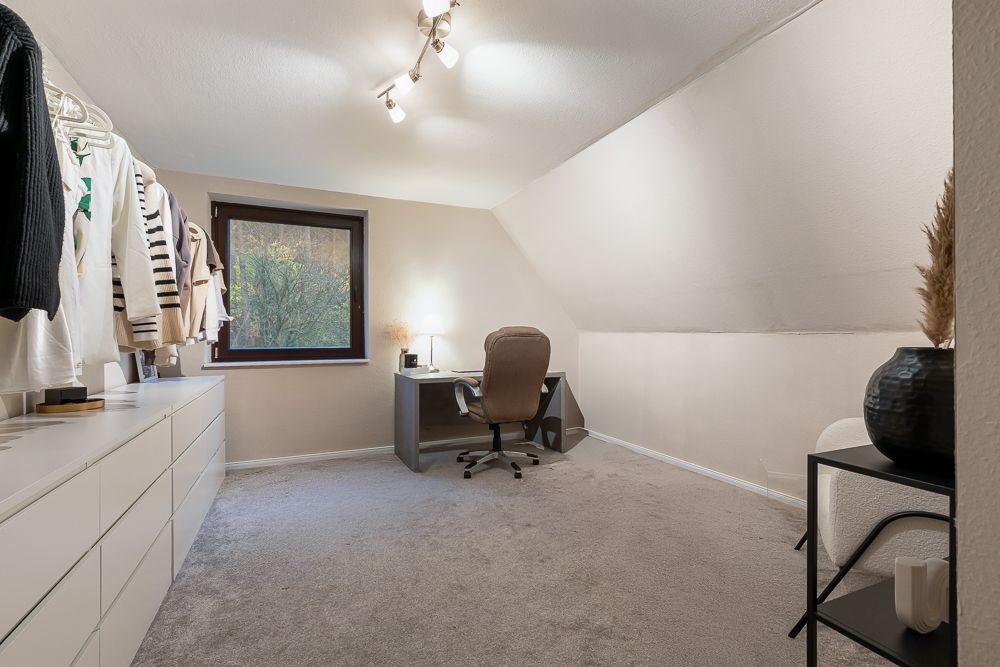 Helles Zimmer mit Schreibtisch, Stuhl, Kleiderstange und Kommode unter Dachschräge.