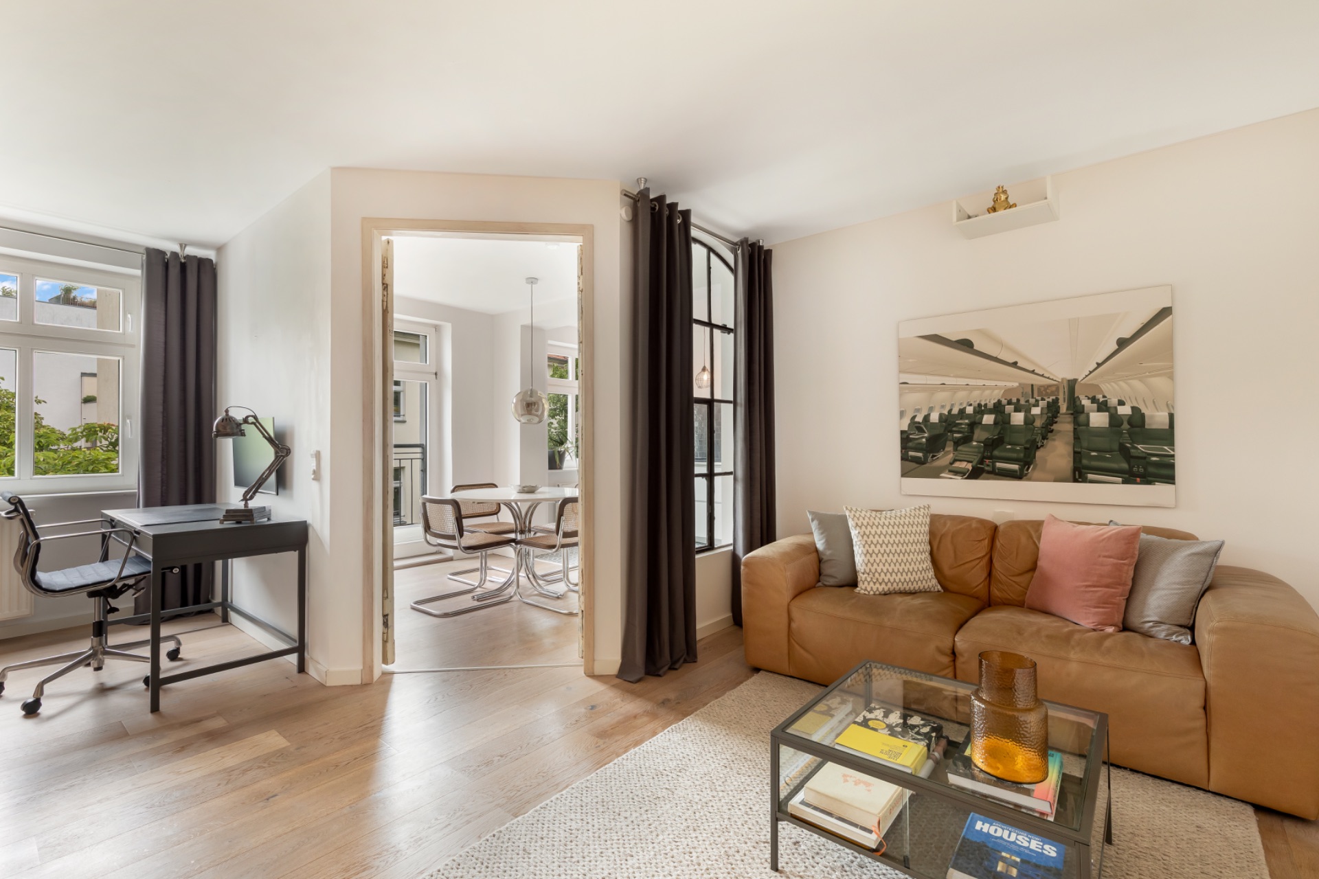 Helles Wohnzimmer mit braunem Sofa, Glastisch, Schreibtisch und Blick in Esszimmer mit rundem Tisch.