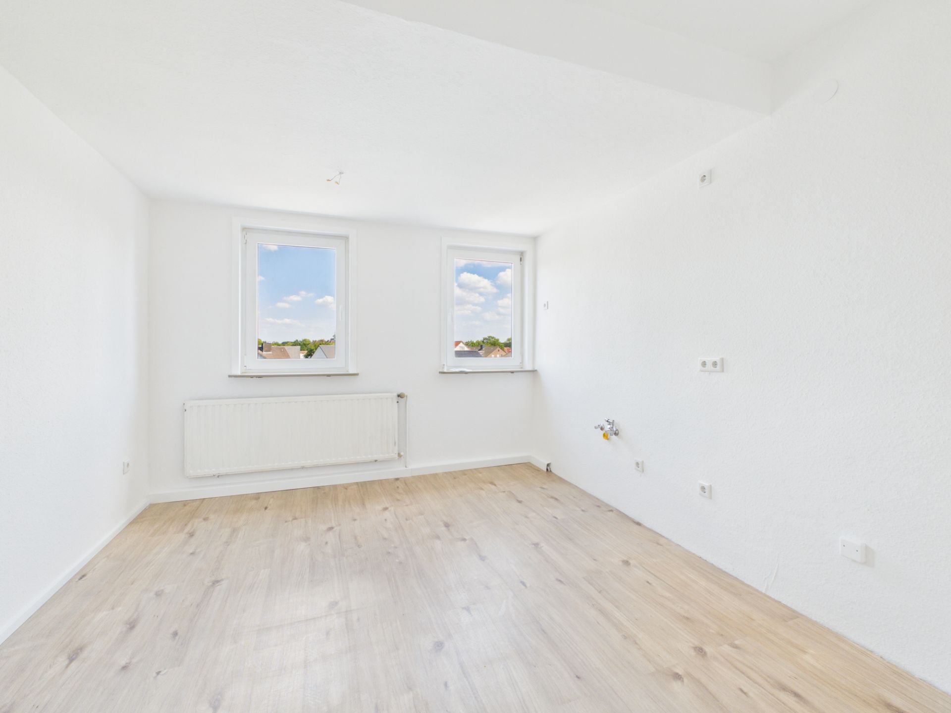 Heller, leerer Raum mit zwei Fenstern, weiß gestrichenen Wänden und hellem Holzfußboden.