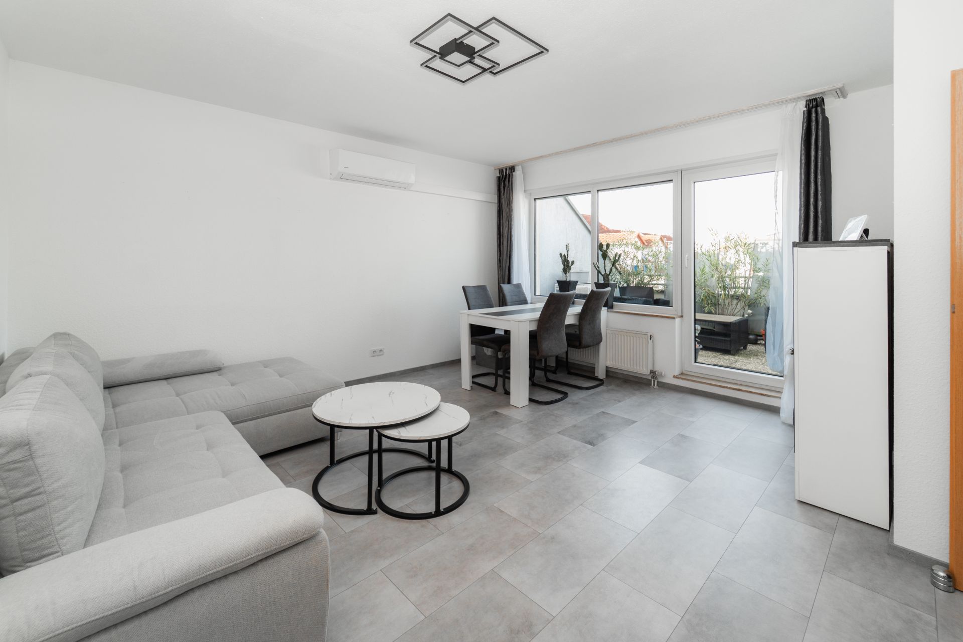 Helles Wohnzimmer mit grauem Sofa, Esstisch, Stühlen, Couchtisch und Zugang zum Balkon.