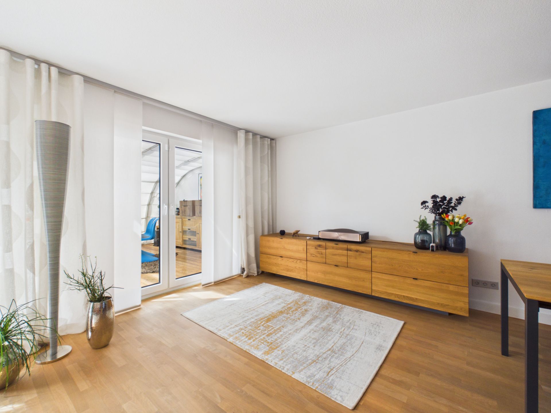 Helles Wohnzimmer mit Holzboden, Teppich, Sideboard, Pflanzen und Glastür zum Balkon.