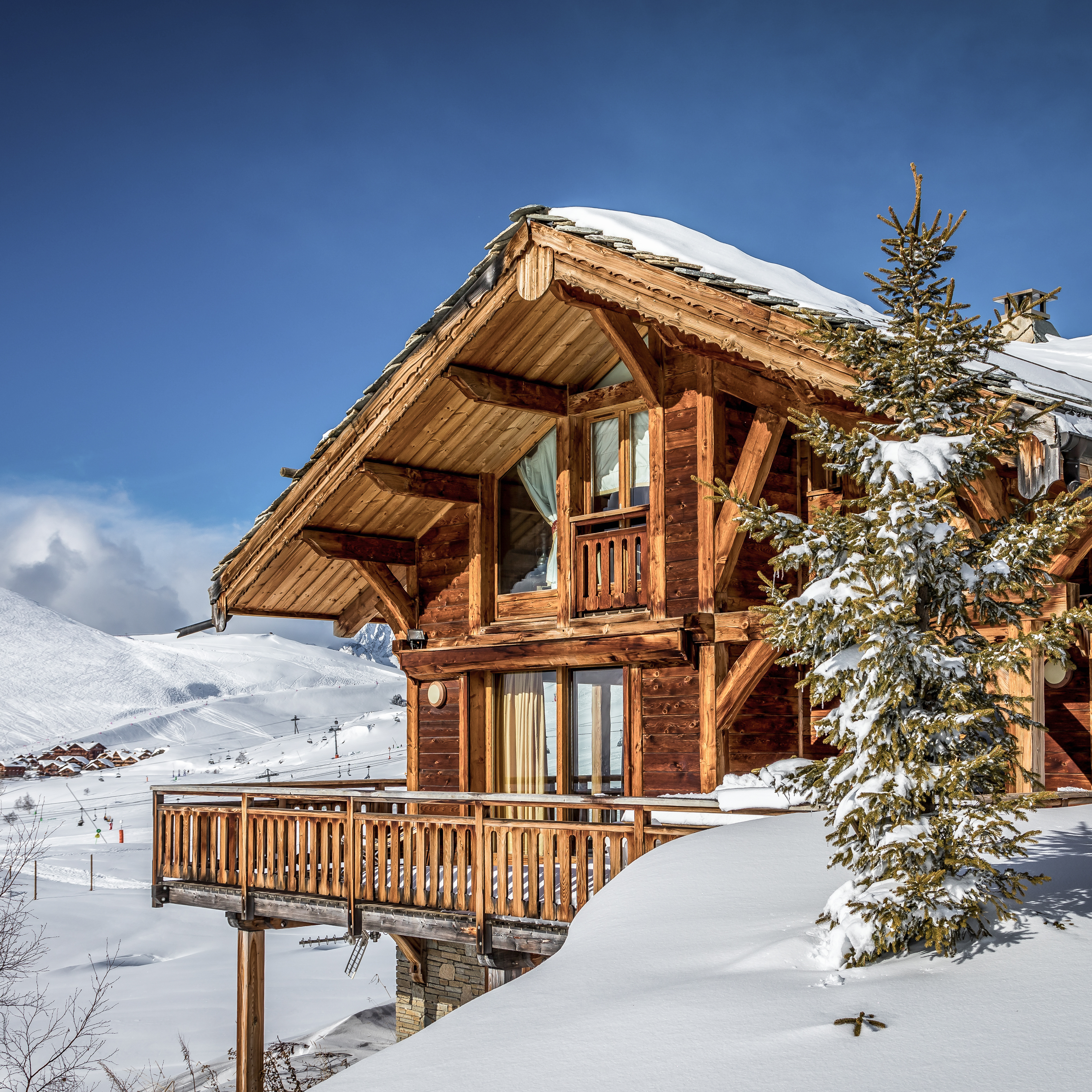 Holzchalet im Schnee mit Balkon und großen Fenstern, Berge im Hintergrund.