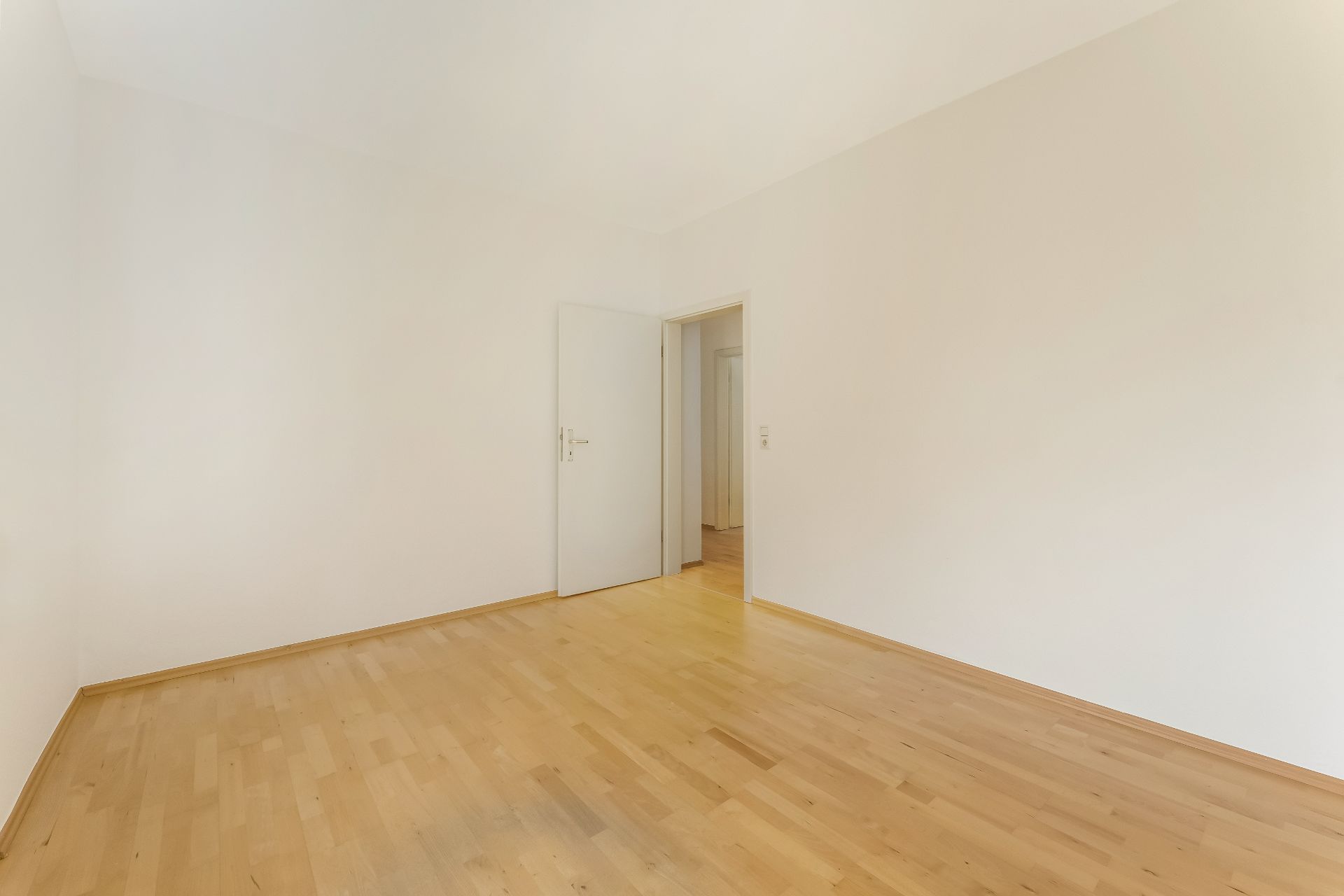 Leerer Raum mit hellem Holzfußboden und weißen Wänden, offene Tür zu einem Flur.