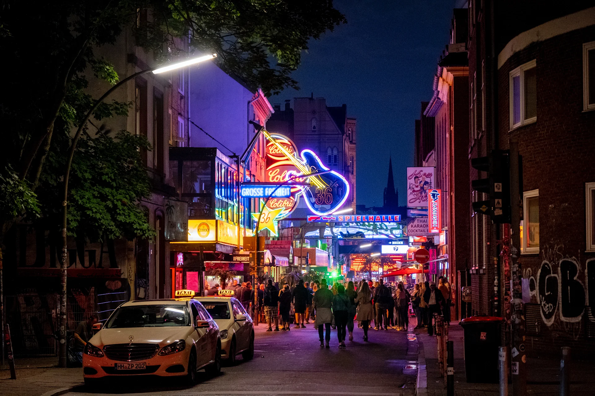 Belebte Straße bei Nacht mit bunten Neonlichtern, Menschen und Taxis am Straßenrand.