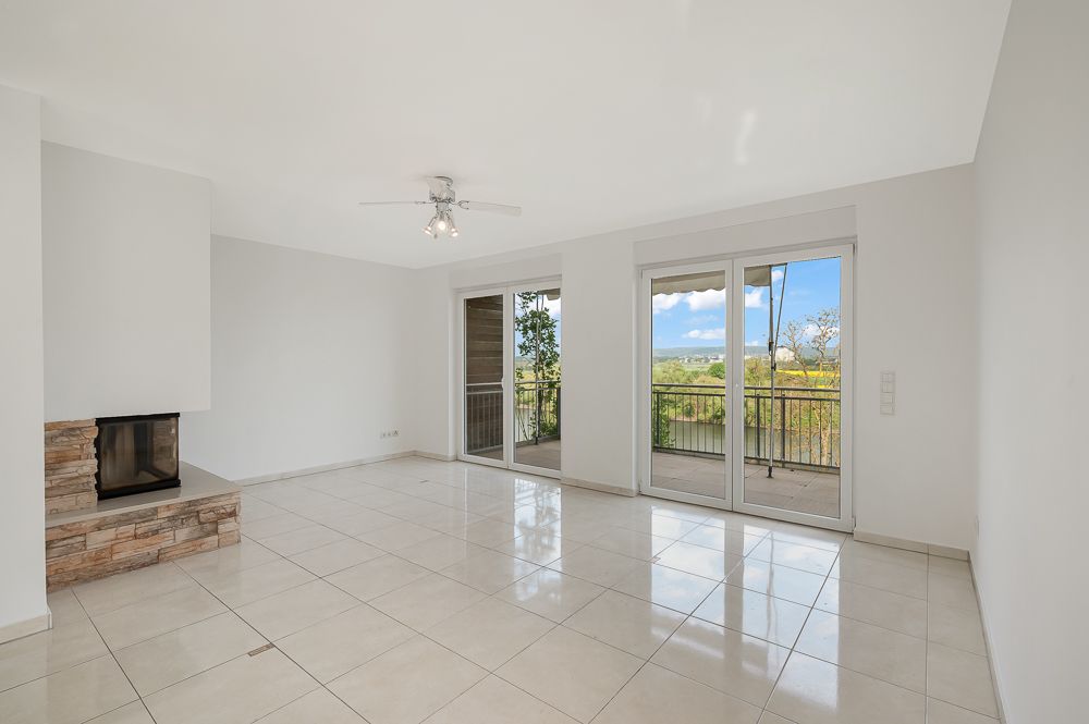 Helles Wohnzimmer mit Kamin, Fliesenboden und großen Fenstern zu einem Balkon mit Ausblick.