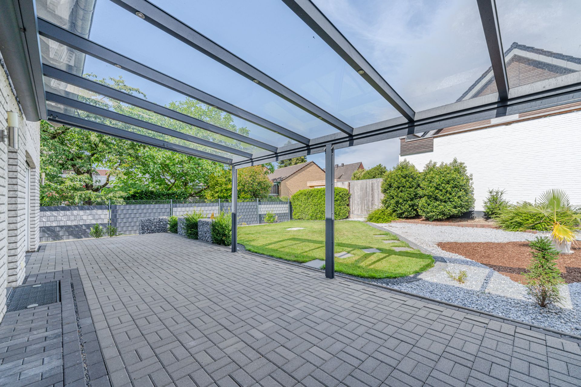 Überdachte Terrasse mit grauen Steinfliesen und Blick auf einen Garten mit Rasen und Sträuchern.