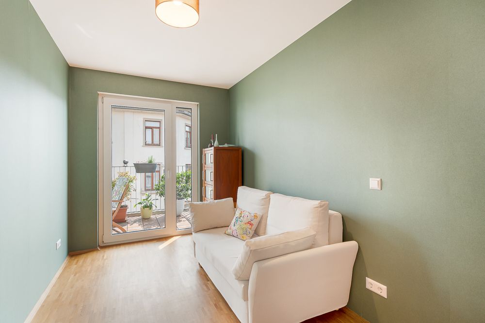 Kleines Zimmer mit grüner Wand, weißem Sofa und Zugang zu einem Balkon mit Pflanzen.
