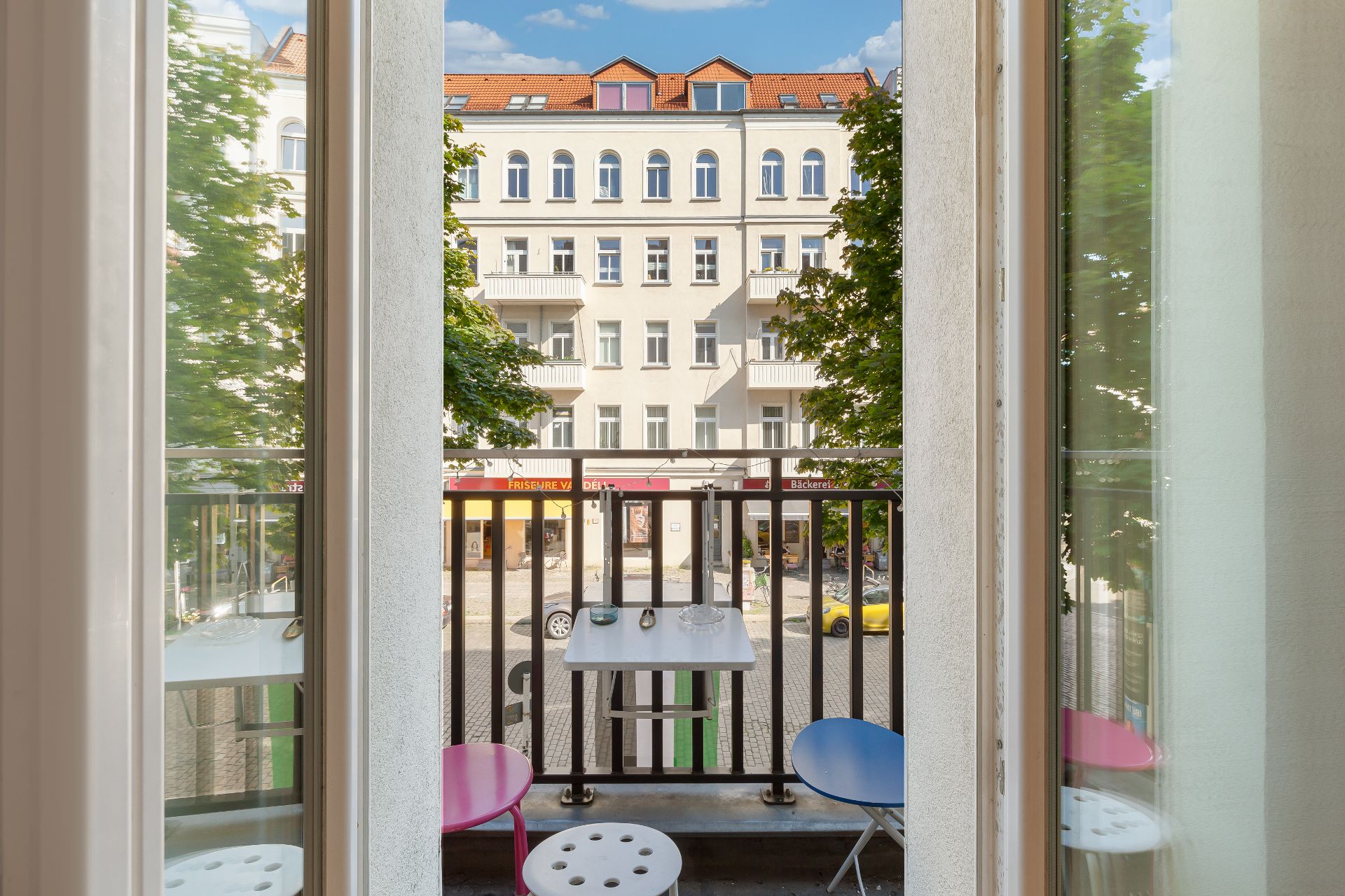 Kleiner Balkon mit Tisch und bunten Hockern, Blick auf Straße und Wohngebäude.