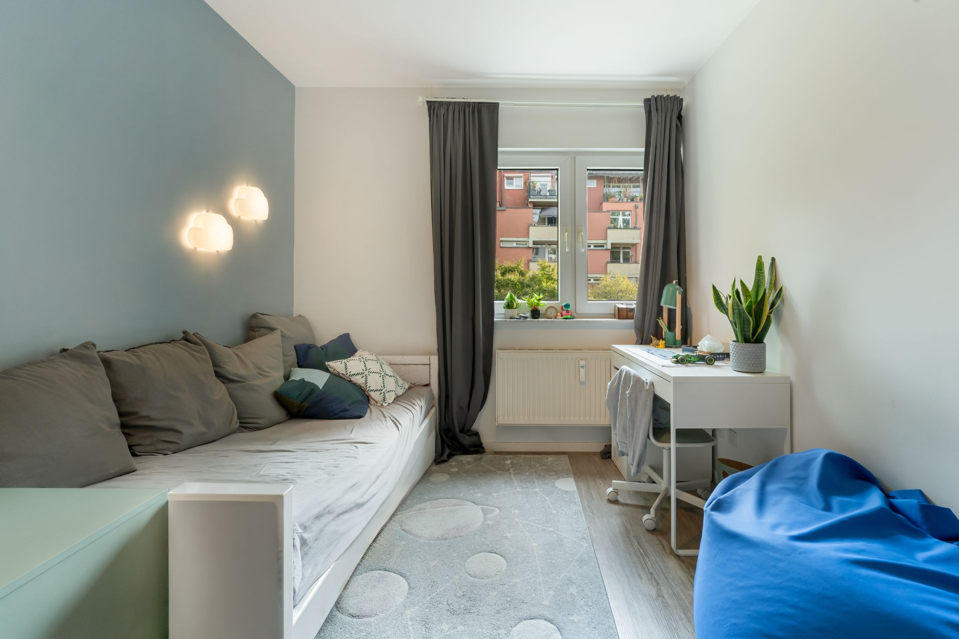 Helles Zimmer mit Sofa, Schreibtisch, Stuhl und Pflanze am Fenster.