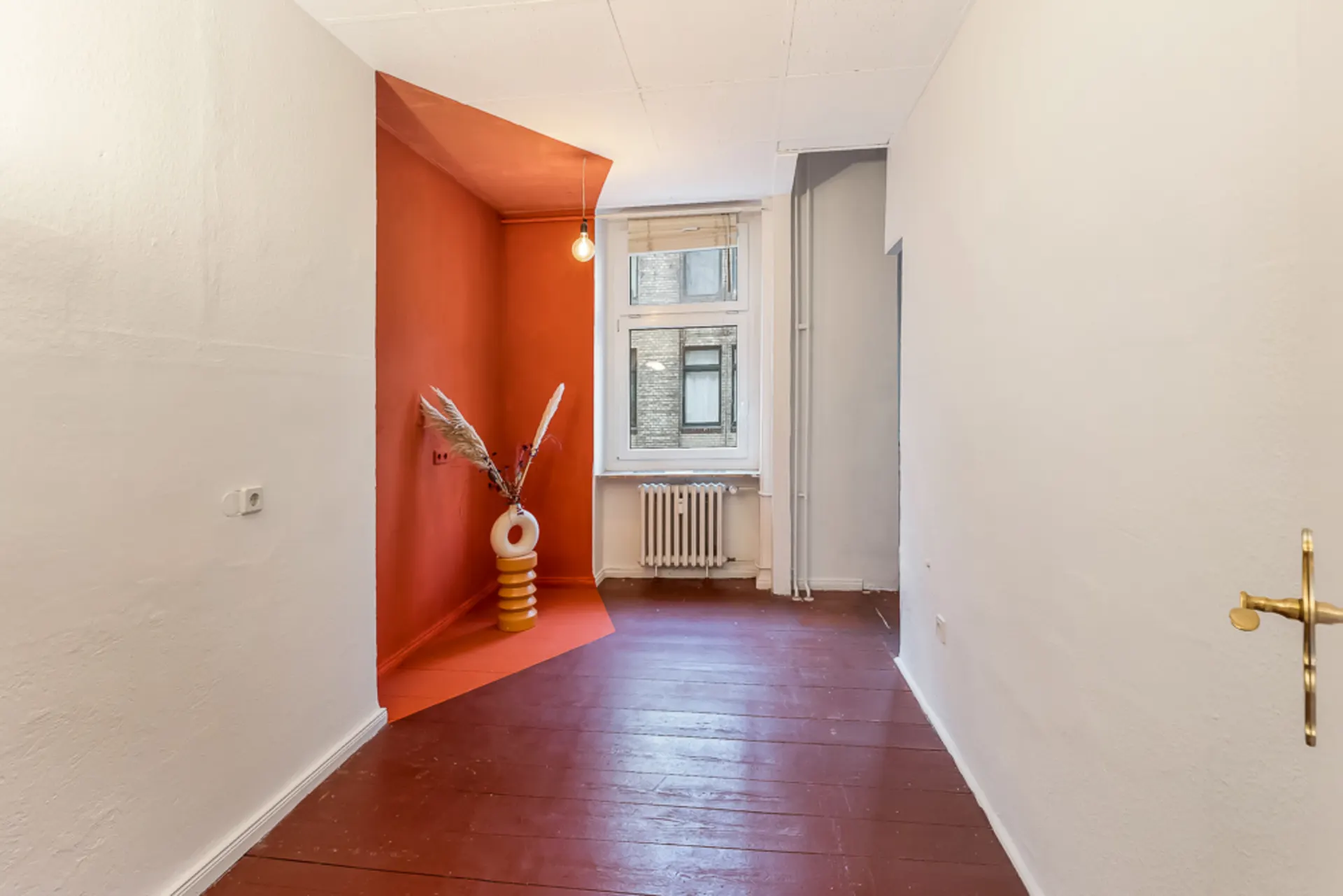 Heller Raum mit rotem Boden, roter Ecke, weißer Vase mit Zweigen und Fenster mit Heizkörper.