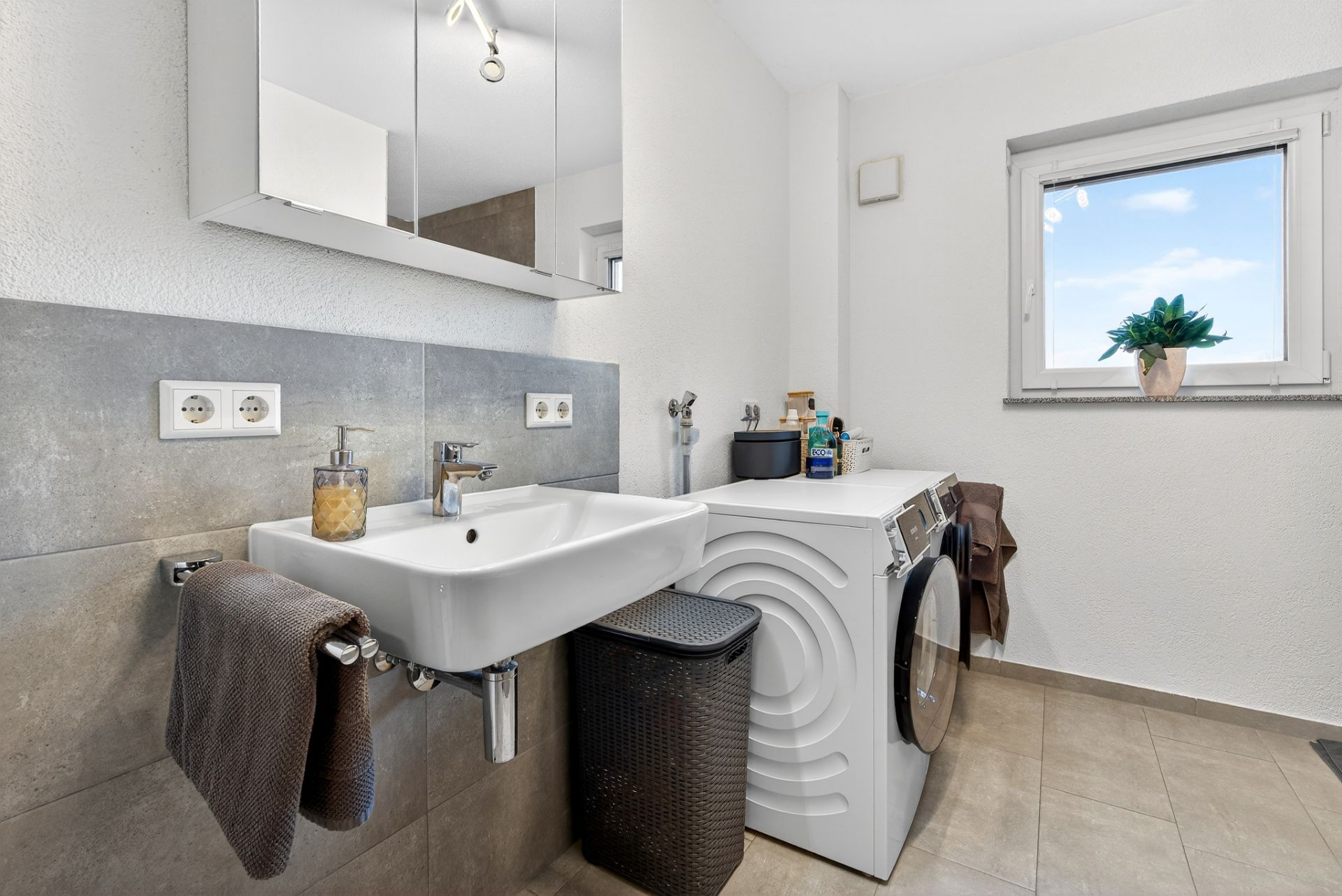 Modernes Badezimmer mit Waschbecken, Spiegel, Waschmaschine und Fenster mit Pflanze.