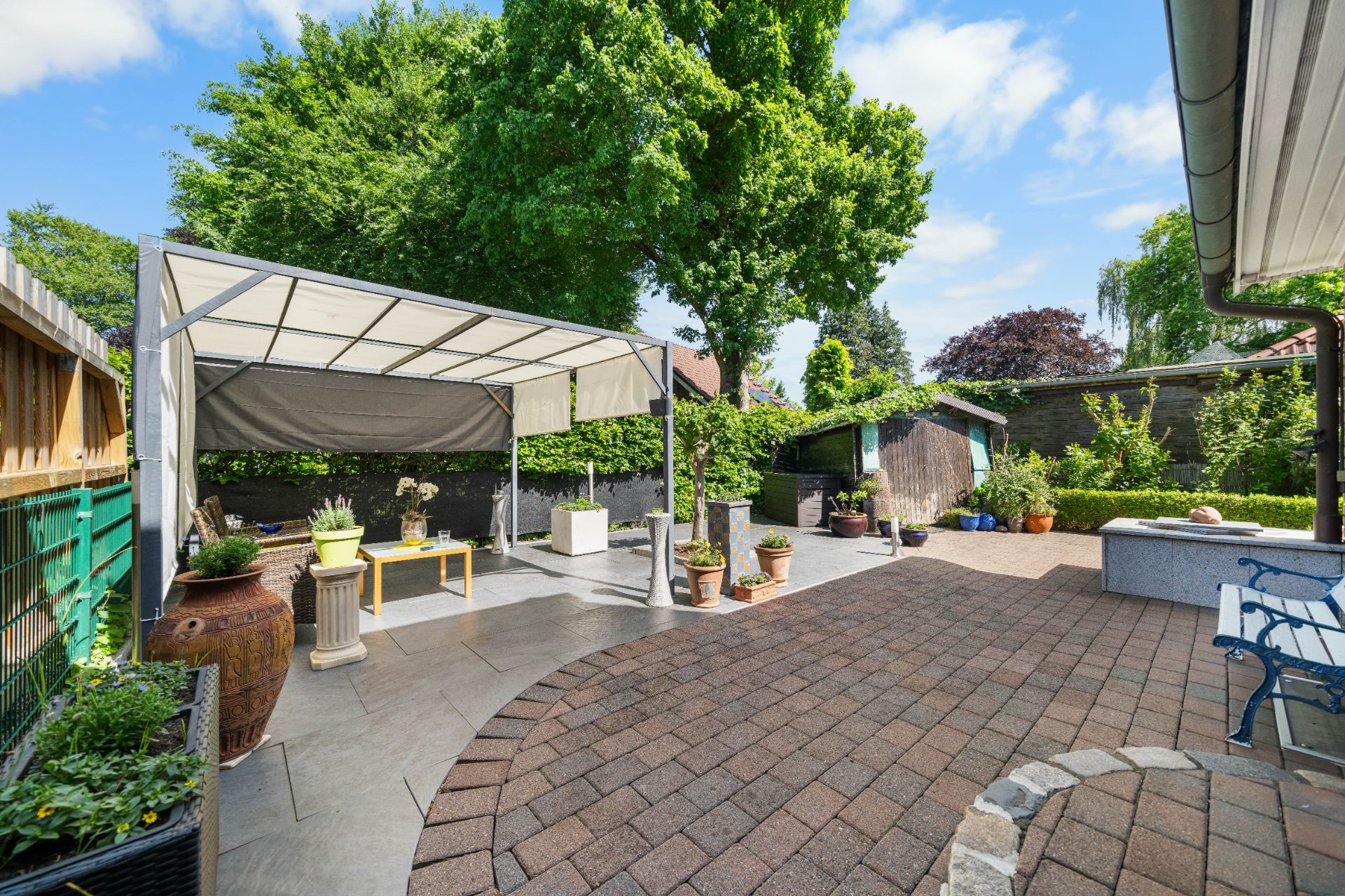Gepflasterte Terrasse mit Sonnenschutz, Sitzbank und vielen Blumentöpfen im Garten.