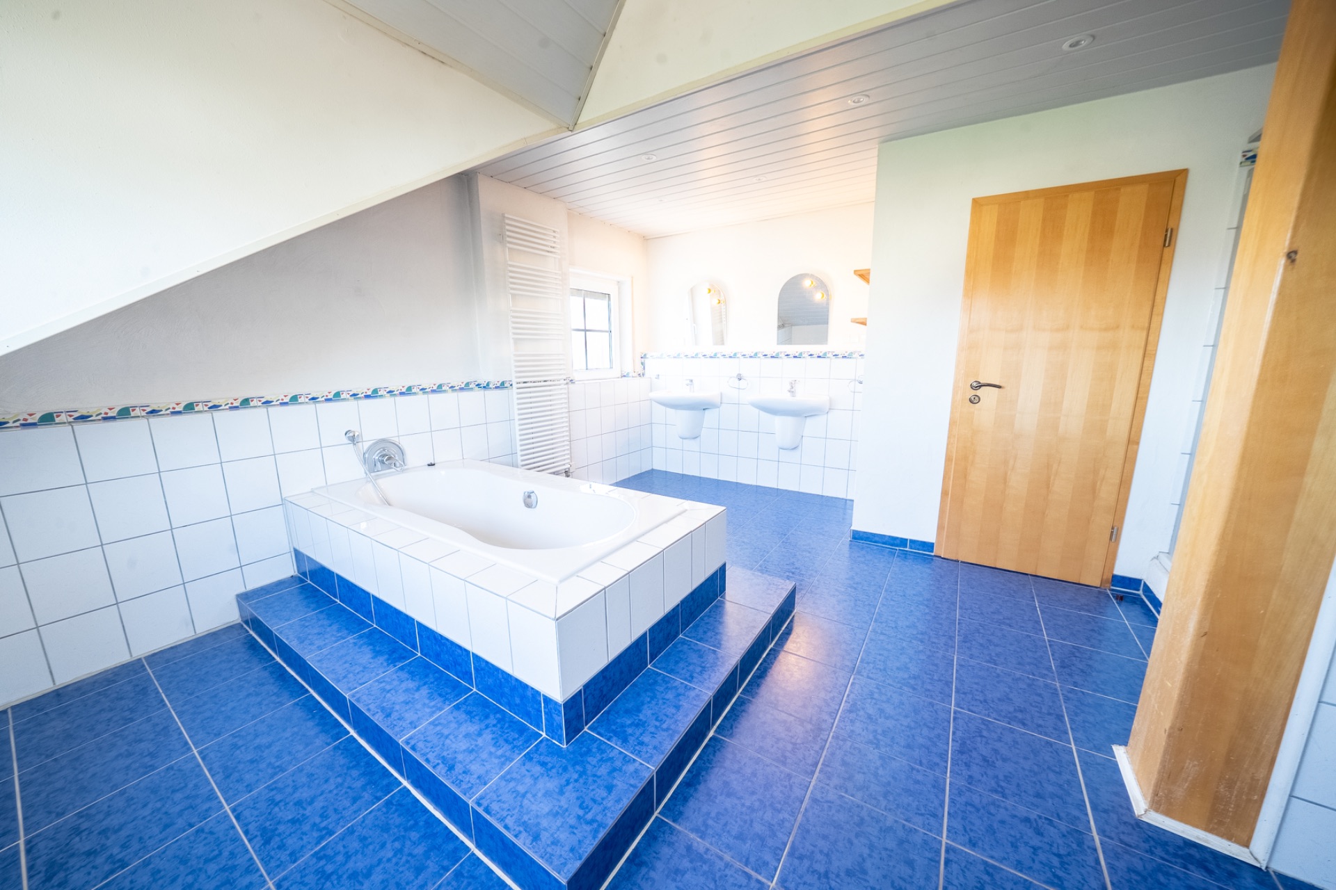 Helles Badezimmer mit Badewanne auf Podest, zwei Waschbecken und blauen Fliesen.