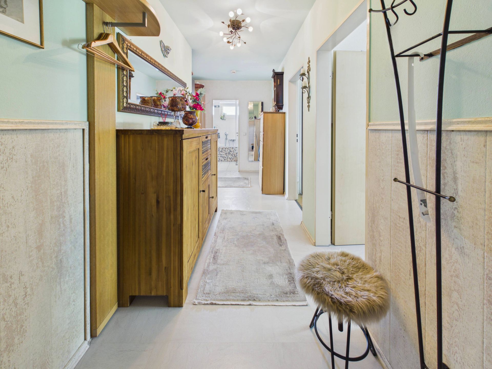 Heller Flur mit Holzkommode, Spiegel, Garderobe und Hocker mit Fellbezug.