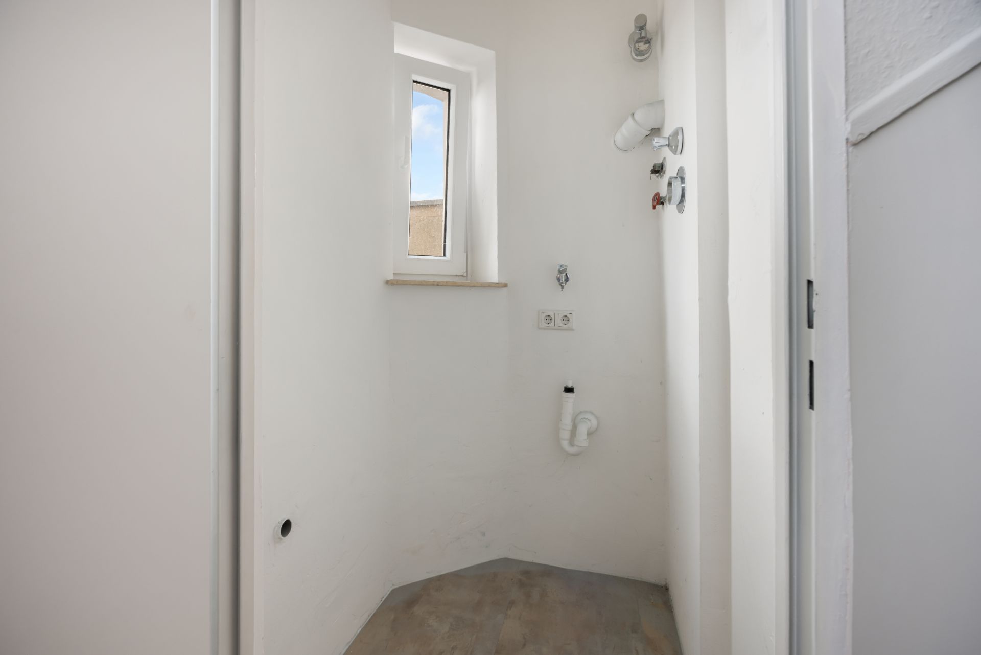 Kleiner Hauswirtschaftsraum mit Fenster, Anschlüssen und hellem Boden.